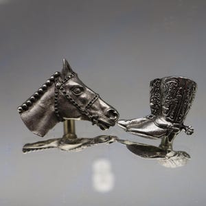 May include: Silver-toned metal horse head and cowboy boot figurines. The horse head features a detailed bridle. The cowboy boot has intricate embossed designs. Both are mounted on small stands, reflecting on the surface below.