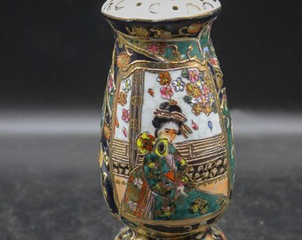 May include: An ornate ceramic salt shaker with a white, perforated top and a flared base. The body features a geisha scene, floral patterns, and gold accents. The color scheme includes green, blue, orange, and gold.
