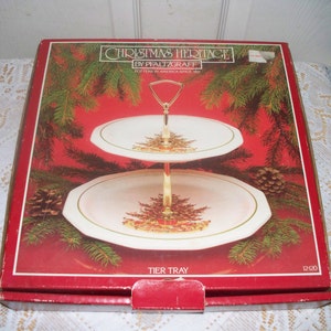 Puede incluir: Una bandeja de servir de dos niveles con un soporte de metal dorado. La bandeja presenta un fondo blanco con un diseño de árbol de Navidad verde y un borde dorado. La caja es roja con las palabras "Christmas Heritage by Pfaltzgraff" y "Tier Tray" impresas en ella.