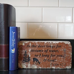 May include: A rustic, rectangular brick with the text "As the deer longs for streams of water, so I long for you, O God! - Psalm 42:1". The brick features a deer and tree silhouette. A blue book and a brown book are to the left.