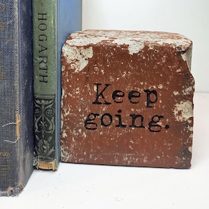 May include: A rustic brick bookend with the words "Keep going" stamped in black. The bookend is placed next to two vintage books with blue covers and a green spine. The brick is a reddish-brown color with white paint.