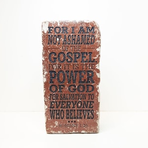 May include: A rectangular, distressed-look decorative block with the text "FOR I AM NOT ASHAMED OF THE GOSPEL, FOR IT IS THE POWER OF GOD FOR SALVATION TO EVERYONE WHO BELIEVES ROMANS 1:16" in black lettering. The block is a reddish-brown color.