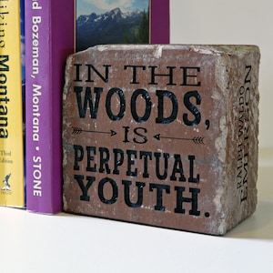 Puede incluir: Un ladrillo r&uacute;stico con la cita "IN THE WOODS, IS PERPETUAL YOUTH" en letras negras. El ladrillo est&aacute; colocado junto a dos libros, uno morado y otro amarillo, con t&iacute;tulos sobre Montana y senderismo.