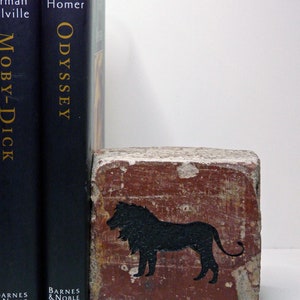 May include: A decorative brick bookend with a black lion silhouette, standing next to two dark blue Barnes & Noble books: "Moby-Dick" and "Odyssey". The bookend has a weathered, reddish-brown appearance.