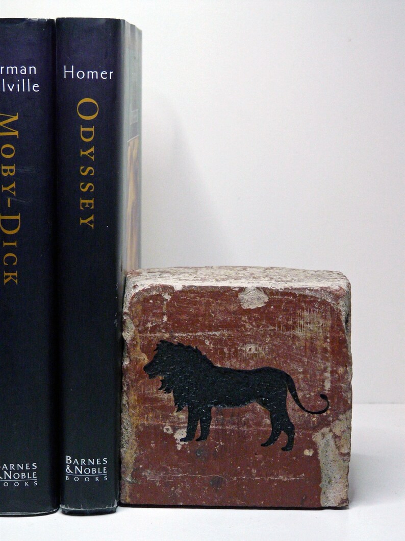 May include: A decorative brick bookend with a black lion silhouette, standing next to two dark blue Barnes & Noble books: "Moby-Dick" and "Odyssey". The bookend has a weathered, reddish-brown appearance.