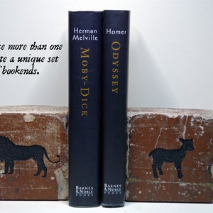 May include: Two bookends made from bricks, each with a black animal silhouette. One features a lion, the other a lamb. The bookends flank two navy blue books, "Moby-Dick" and "Odyssey."