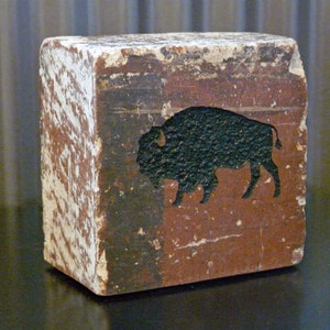 May include: A weathered, rectangular wooden block with a painted bison silhouette. The block has a distressed, aged appearance with white and brown paint. The bison is black, set against a reddish-brown background. The block is a decorative item.