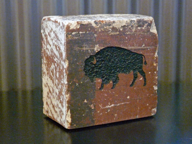 May include: A weathered, rectangular wooden block with a painted bison silhouette. The block has a distressed, aged appearance with white and brown paint. The bison is black, set against a reddish-brown background. The block is a decorative item.