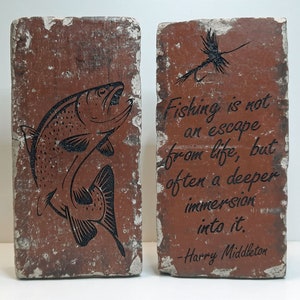 May include: Two rectangular, weathered, reddish-brown decorative bricks. One brick features a black illustration of a leaping fish. The other brick has a fishing fly illustration and the quote: "Fishing is not an escape from life, but often a deeper immersion into it."