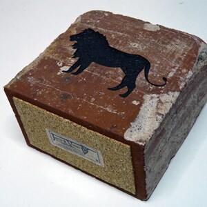 May include: A rectangular, brick-like object with a black lion silhouette on top. The object is primarily reddish-brown with a lighter, textured border. A label is visible on the side. The overall aesthetic suggests a vintage or rustic style.