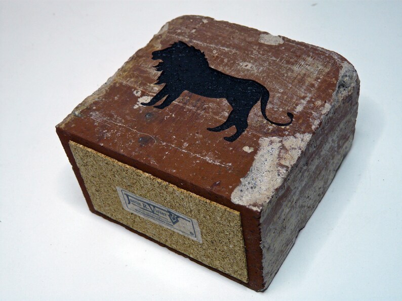 May include: A rectangular, brick-like object with a black lion silhouette on top. The object is primarily reddish-brown with a lighter, textured border. A label is visible on the side. The overall aesthetic suggests a vintage or rustic style.