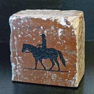 K&ouml;nnte beinhalten: Rechteckiger Steinblock mit einer schwarzen Silhouette eines Reiters auf einem Pferd. Der Stein hat einen r&ouml;tlich-braunen Farbton mit rauer Oberfl&auml;che und wei&szlig;en Kanten. Western-Thema.