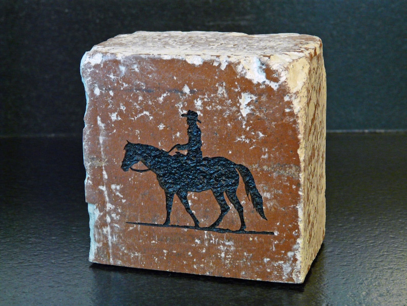 K&ouml;nnte beinhalten: Rechteckiger Steinblock mit einer schwarzen Silhouette eines Reiters auf einem Pferd. Der Stein hat einen r&ouml;tlich-braunen Farbton mit rauer Oberfl&auml;che und wei&szlig;en Kanten. Western-Thema.