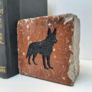 May include: A rustic, rectangular bookend made of reddish-brown brick. A black silhouette of a dog is carved into the brick. The bookend is next to three black books with gold lettering.