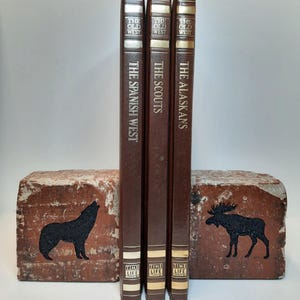 Op de afbeelding: Drie vintage boeken uit de Time-Life Old West-serie, getiteld "The Spanish West", "The Scouts" en "The Alaskans". De boeken zijn bruin met gouden letters. Ze worden vastgehouden door twee bakstenen boekensteunen met zwarte dierensilhouetten.