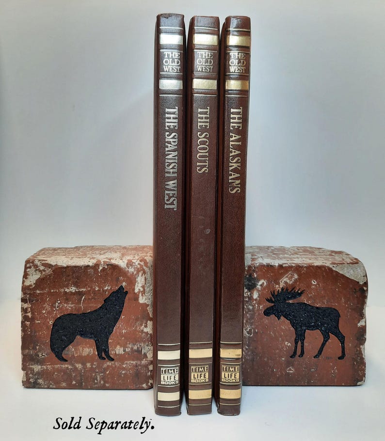 Op de afbeelding: Drie vintage boeken uit de Time-Life Old West-serie, getiteld "The Spanish West", "The Scouts" en "The Alaskans". De boeken zijn bruin met gouden letters. Ze worden vastgehouden door twee bakstenen boekensteunen met zwarte dierensilhouetten.