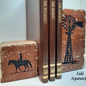 K&ouml;nnte beinhalten: Rustikale Buchst&uuml;tzen mit Western-Thema. Eine Buchst&uuml;tze zeigt eine schwarze Silhouette eines Cowboys auf einem Pferd, die andere eine Windm&uuml;hle. Drei braune Hardcover-B&uuml;cher mit goldfarbenen Buchstaben sind zwischen den Buchst&uuml;tzen aufgestellt. Die B&uuml;cher tragen die Titel "The Rivermen", "The Miners" und "The Cowboys".