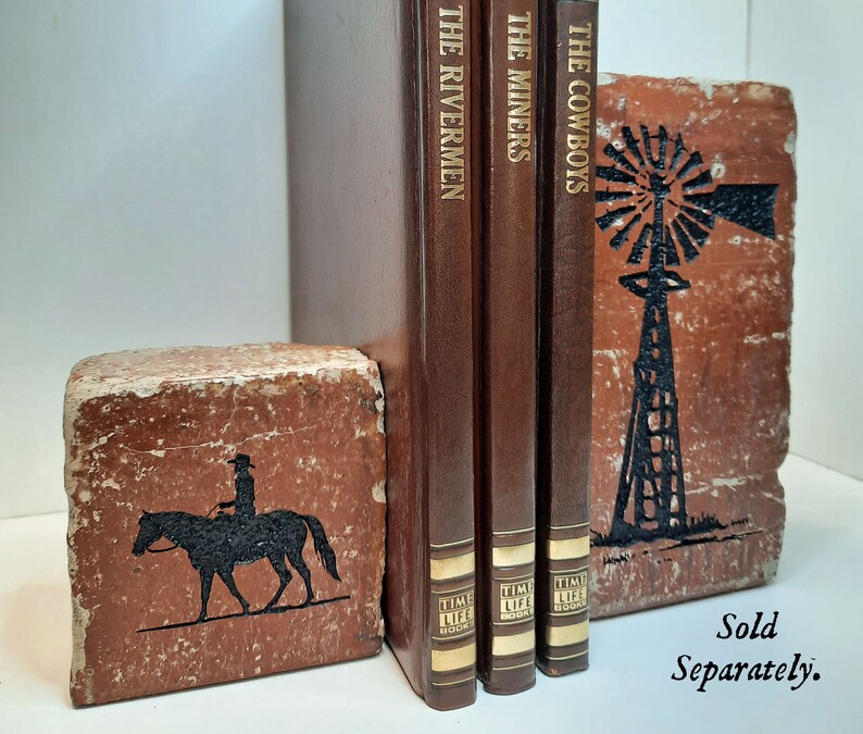 K&ouml;nnte beinhalten: Rustikale Buchst&uuml;tzen mit Western-Thema. Eine Buchst&uuml;tze zeigt eine schwarze Silhouette eines Cowboys auf einem Pferd, die andere eine Windm&uuml;hle. Drei braune Hardcover-B&uuml;cher mit goldfarbenen Buchstaben sind zwischen den Buchst&uuml;tzen aufgestellt. Die B&uuml;cher tragen die Titel "The Rivermen", "The Miners" und "The Cowboys".