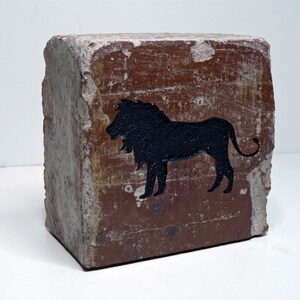 May include: A weathered, rectangular brick with a rough, textured surface. A black silhouette of a lion is painted on the brick's face. The brick has a reddish-brown hue with areas of exposed, lighter-colored aggregate.