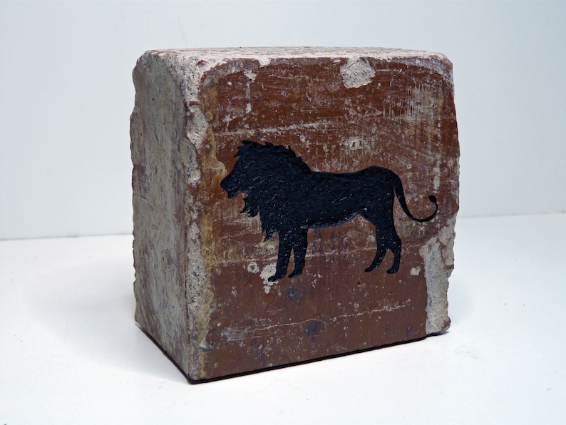 May include: A weathered, rectangular brick with a rough, textured surface. A black silhouette of a lion is painted on the brick's face. The brick has a reddish-brown hue with areas of exposed, lighter-colored aggregate.