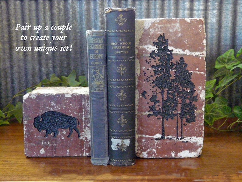 May include: A set of rustic bookends made from repurposed bricks. One bookend features a black bison silhouette, the other a black tree design. Two antique books stand between the bookends. The text on the image reads: "Pair up a couple to create your own unique set!"