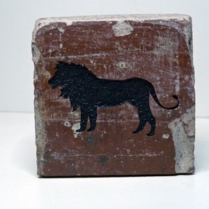 May include: A weathered, rectangular brick with a dark brown surface. A black silhouette of a lion is painted on the front. The brick has chipped edges and a distressed appearance, suggesting age or wear.