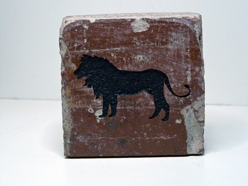 May include: A weathered, rectangular brick with a dark brown surface. A black silhouette of a lion is painted on the front. The brick has chipped edges and a distressed appearance, suggesting age or wear.