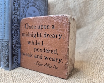 Puede incluir: Un ladrillo rectangular de terracota con una textura rugosa y desgastada que presenta el texto impreso en negro: Once upon a midnight dreary, while I pondered, weak and weary. - Edgar Allan Poe. El ladrillo tiene un frente rojo-marrón.