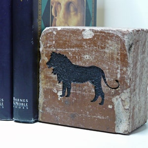 May include: A rectangular, brown bookend with a black lion silhouette. The bookend has a distressed, aged appearance. Three navy blue books are visible to the left of the bookend, with the words "Barnes & Noble Books" printed on the spines.
