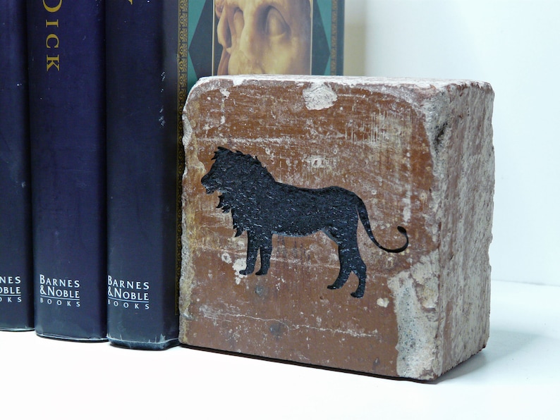 May include: A rectangular, brown bookend with a black lion silhouette. The bookend has a distressed, aged appearance. Three navy blue books are visible to the left of the bookend, with the words "Barnes & Noble Books" printed on the spines.