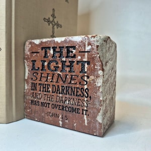 May include: A rectangular, weathered brick with the text "THE LIGHT SHINES IN THE DARKNESS, AND THE DARKNESS HAS NOT OVERCOME IT. JOHN 1:5" in black. The brick is reddish-brown with white accents.