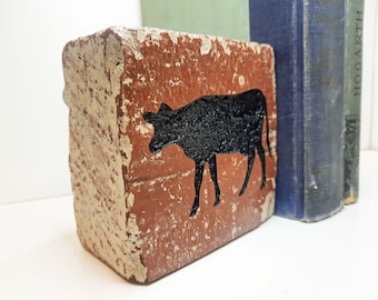 May include: A rustic, rectangular brick bookend featuring a black cow silhouette on a red-brown background. The bookend has a distressed, textured appearance. Vintage books are visible in the background.