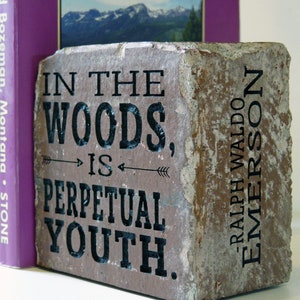 Puede incluir: Un sujetalibros de piedra rectangular con la cita "IN THE WOODS, IS PERPETUAL YOUTH" y "RALPH WALDO EMERSON" en letras negras. El sujetalibros es una mezcla de tonos marrones y beige, con un libro morado visible detr&aacute;s.