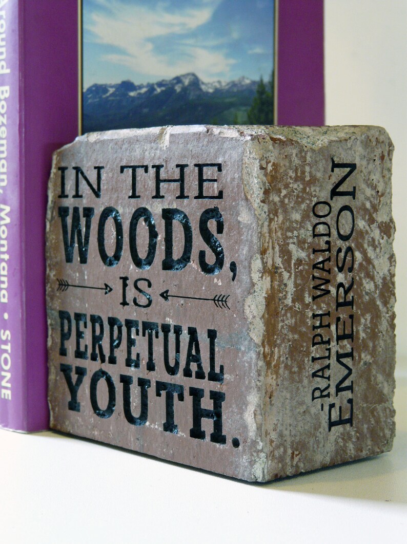 Puede incluir: Un sujetalibros de piedra rectangular con la cita "IN THE WOODS, IS PERPETUAL YOUTH" y "RALPH WALDO EMERSON" en letras negras. El sujetalibros es una mezcla de tonos marrones y beige, con un libro morado visible detr&aacute;s.