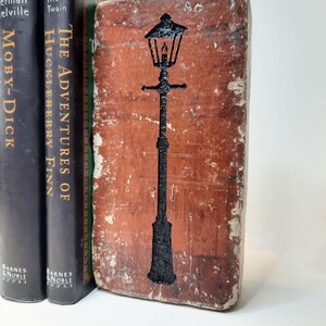 May include: A weathered, brick-effect bookend featuring a black outline of a traditional street lamp. It stands beside two classic novels: "Moby-Dick" and "The Adventures of Huckleberry Finn".