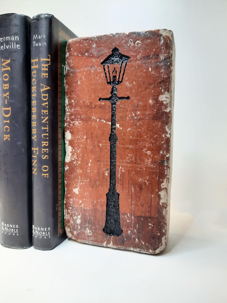 May include: A weathered, brick-effect bookend featuring a black outline of a traditional street lamp. It stands beside two classic novels: "Moby-Dick" and "The Adventures of Huckleberry Finn".