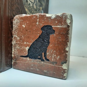 May include: A rustic, rectangular brick featuring a black silhouette of a dog. The brick has a weathered, reddish-brown surface with a distressed, textured appearance. The dog is sitting, and the brick is placed near brown books.