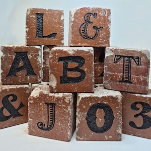 May include: A collection of brown, distressed wooden blocks, each displaying a black letter or symbol. The letters include "L", "A", "B", "T", "J", "O", and the number "3", along with the ampersand symbol. The blocks have a rustic, vintage aesthetic.