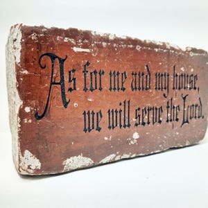 May include: A rectangular, weathered brick with the inscription "As for me and my house, we will serve the Lord" in black gothic lettering. The brick is primarily reddish-brown with white accents.