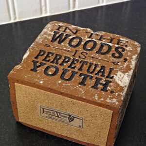 Puede incluir: Ladrillo rectangular desgastado con la cita "IN THE WOODS IS PERPETUAL YOUTH" en letras negras. El ladrillo tiene una base de corcho y una peque&ntilde;a etiqueta con un nombre y un sitio web.