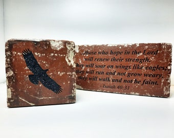 May include: Two rectangular, weathered, red-brown bricks. One features a black eagle silhouette. The other displays text from Isaiah 40:31: "...those who hope in the Lord will renew their strength..." The bricks have a rustic, aged appearance.