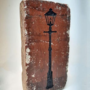 May include: A weathered brick featuring a black silhouette of a traditional street lamp. The brick is a reddish-brown hue, with white paint chipping around the edges. The lamp design includes a lantern and a tall, slender post.