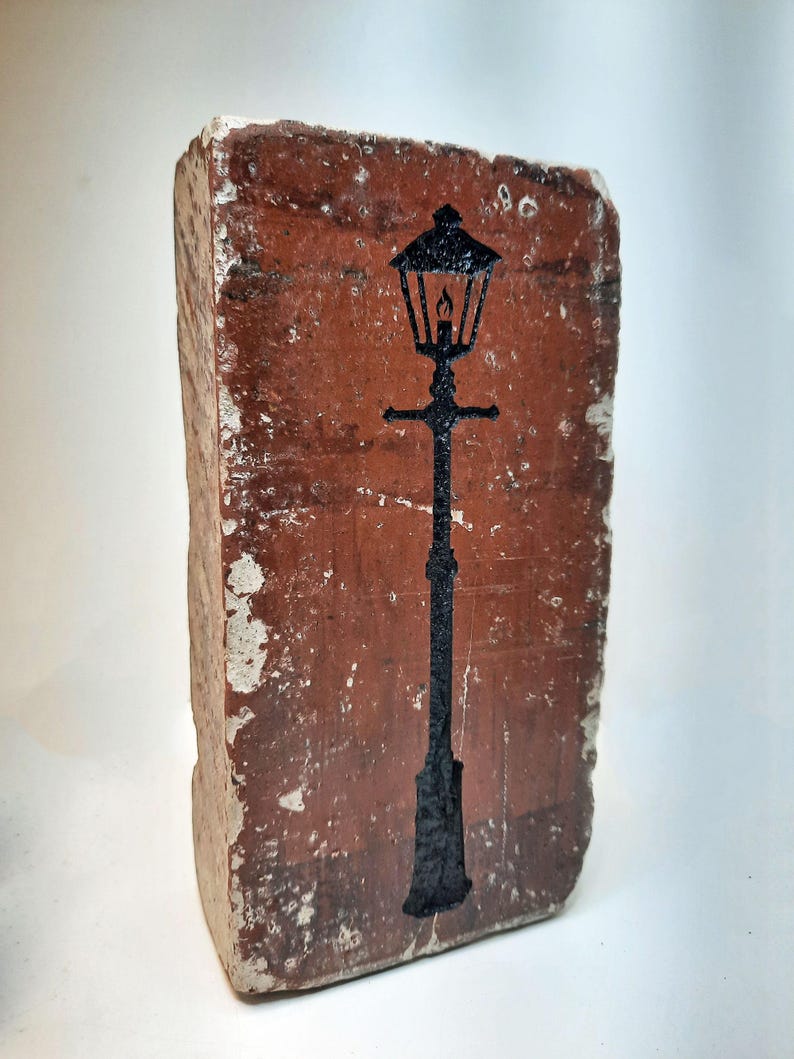May include: A weathered brick featuring a black silhouette of a traditional street lamp. The brick is a reddish-brown hue, with white paint chipping around the edges. The lamp design includes a lantern and a tall, slender post.