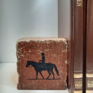 K&ouml;nnte beinhalten: Ein rustikaler, rechteckiger Buchst&uuml;tze mit einer verwitterten, rotbraunen Oberfl&auml;che. Auf der Vorderseite befindet sich eine schwarze Silhouette einer Person auf einem Pferd. Zwei braune Hardcover-B&uuml;cher mit goldener Schrift sind sichtbar.