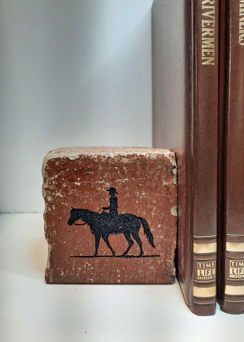 K&ouml;nnte beinhalten: Ein rustikaler, rechteckiger Buchst&uuml;tze mit einer verwitterten, rotbraunen Oberfl&auml;che. Auf der Vorderseite befindet sich eine schwarze Silhouette einer Person auf einem Pferd. Zwei braune Hardcover-B&uuml;cher mit goldener Schrift sind sichtbar.