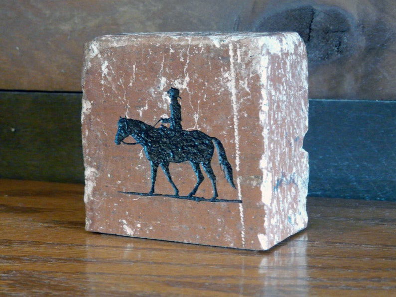 K&ouml;nnte beinhalten: Ein rechteckiger, verwitterter Ziegelstein mit rotbrauner Farbe zeigt eine schwarze Silhouette eines Reiters auf einem Pferd. Der Ziegel hat wei&szlig;e Akzente an den Kanten. Klassische Darstellung einer Reitszene.