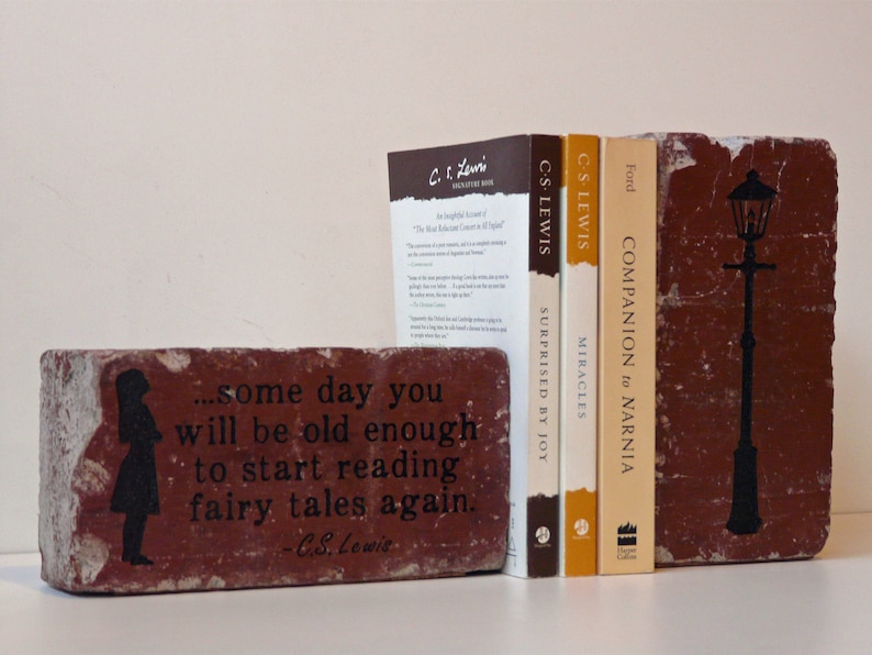 May include: A set of decorative brick bookends with a literary theme. One bookend features a silhouette of a girl and a quote by C.S. Lewis. The other bookend has a black lamppost design. Three books are between the bookends.