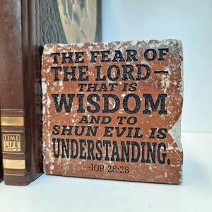 May include: A weathered, rectangular plaque with the text "THE FEAR OF THE LORD - THAT IS WISDOM AND TO SHUN EVIL IS UNDERSTANDING. - JOB 28:28" in black lettering. The plaque is brown with white accents, and is next to a row of brown books.