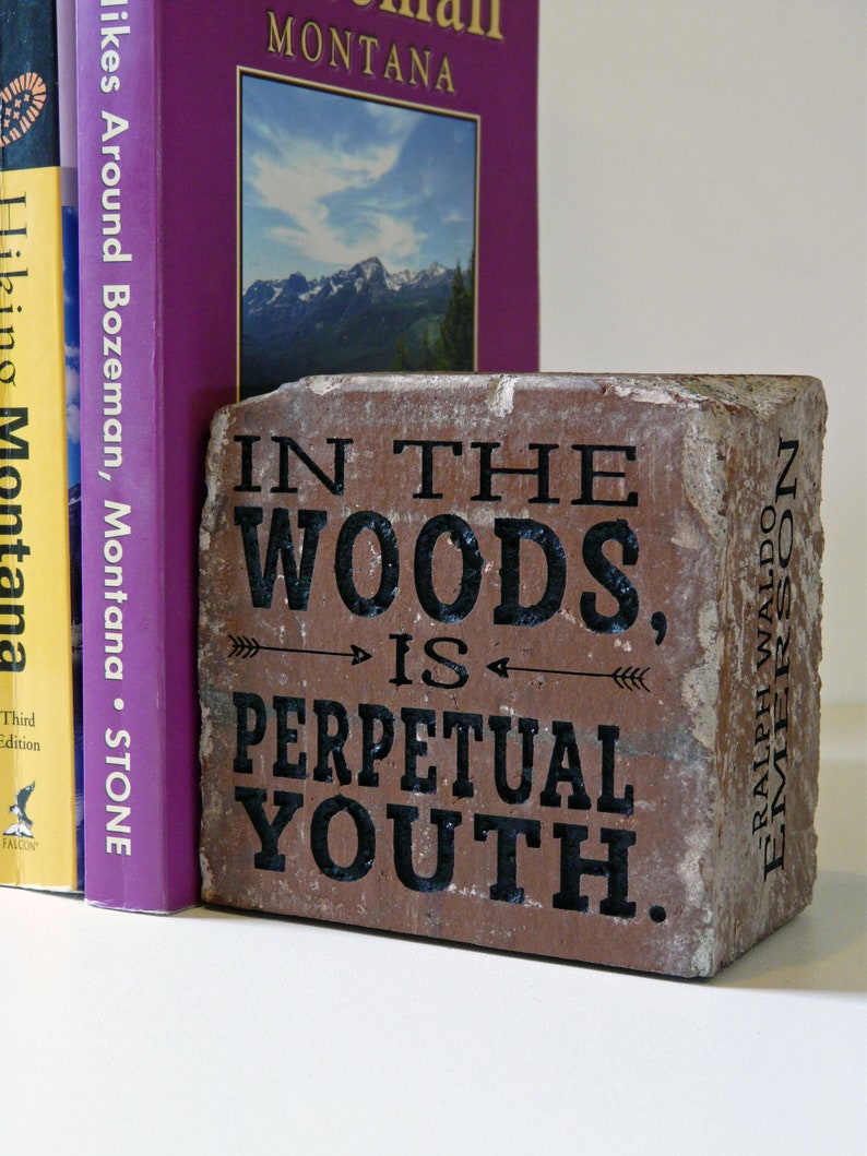 Puede incluir: Un ladrillo r&uacute;stico con la cita "IN THE WOODS, IS PERPETUAL YOUTH" en letras negras. El ladrillo est&aacute; colocado junto a dos libros, uno morado y otro amarillo, con t&iacute;tulos sobre Montana y senderismo.