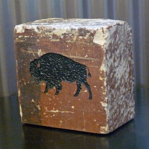 May include: A weathered, rectangular block with a brown base and a black bison silhouette. The block has a distressed, aged appearance with white paint chipping off the edges, giving it a rustic aesthetic.